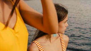 Two women in swimsuits stand on rocks at the water's edge. One clutches a straw hat as she gazes out over the Mediterranean