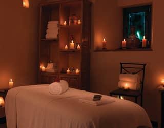 Banks of candles glow on the shelves of a therapy room in low light, around a treatment bed with rolled and folded towels.