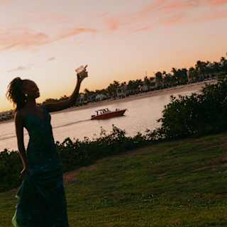 A woman holding up a drink is silhouetted against a pink sunset sky that tints the bay behind, where a small boat cruises by.