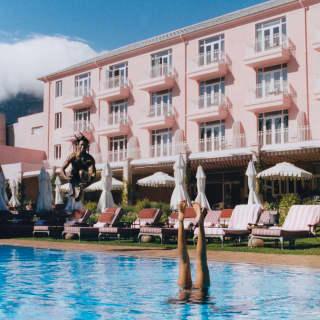 Action shot as man cannon-jumps into the pool where a swimmer performs an underwater handstand, overlooked by the pink hotel.