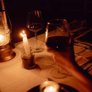In the darkness of the bar at night, a guest raises a glass of red wine at a table illuminated by two candles and a lamp.