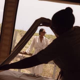 A woman in a white robe and shades leans on the deck rails at the spa as a staff member in the foreground arranges towels.