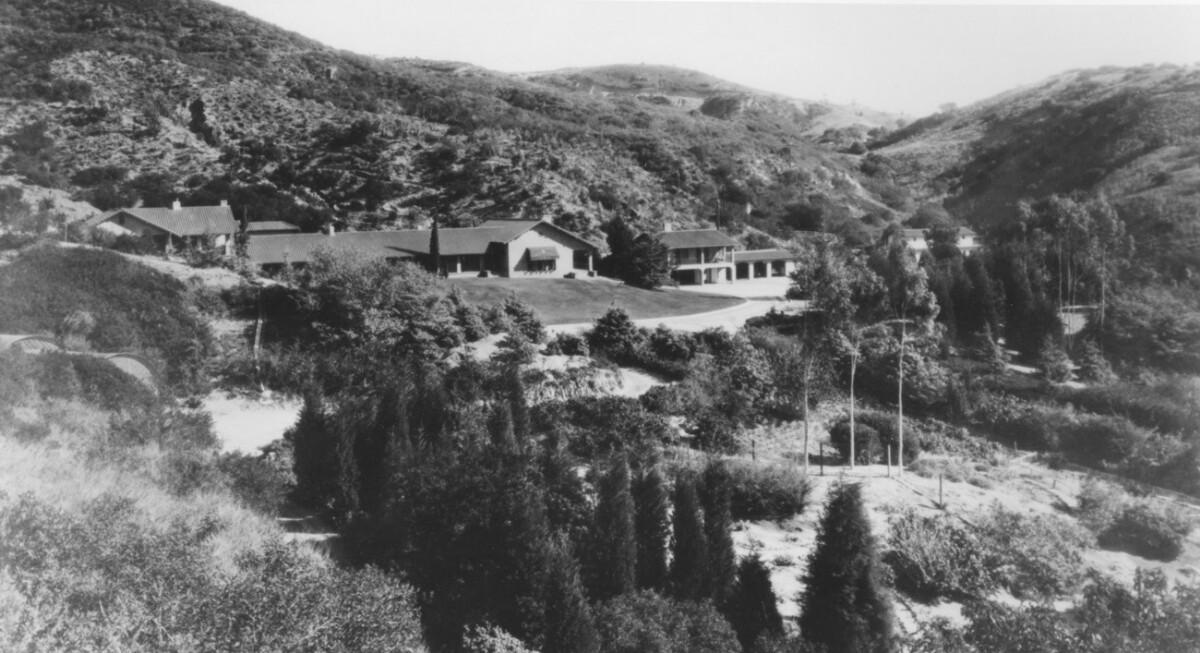 Black and white photo of a house nestled in natural undeveloped hills with brush and trees.