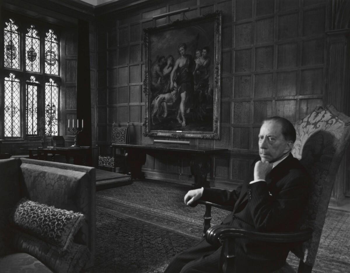 Black and white photo of J. Paul Getty sitting in a tall chair in a grandiose room with wood panel walls, cathedral windows, and complex rugs and furniture
