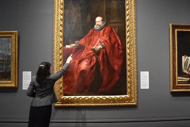 A large portrait of a man in a vivid red robe with a red frame hangs in a gallery setting, while a woman in the lower left of the frame points up at the man.