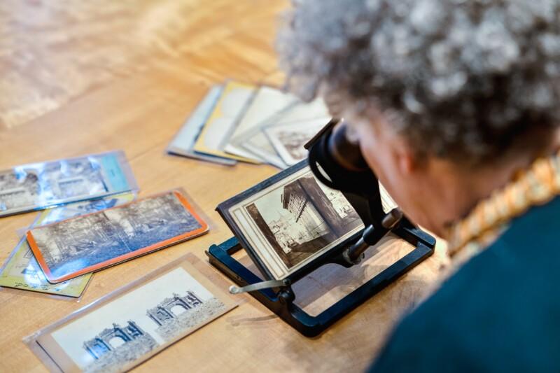 A person seated before a large wooden table looks at a black-and-white stereograph of a streetscape through a modern stereoscope, while other stereographs are scattered on the table.