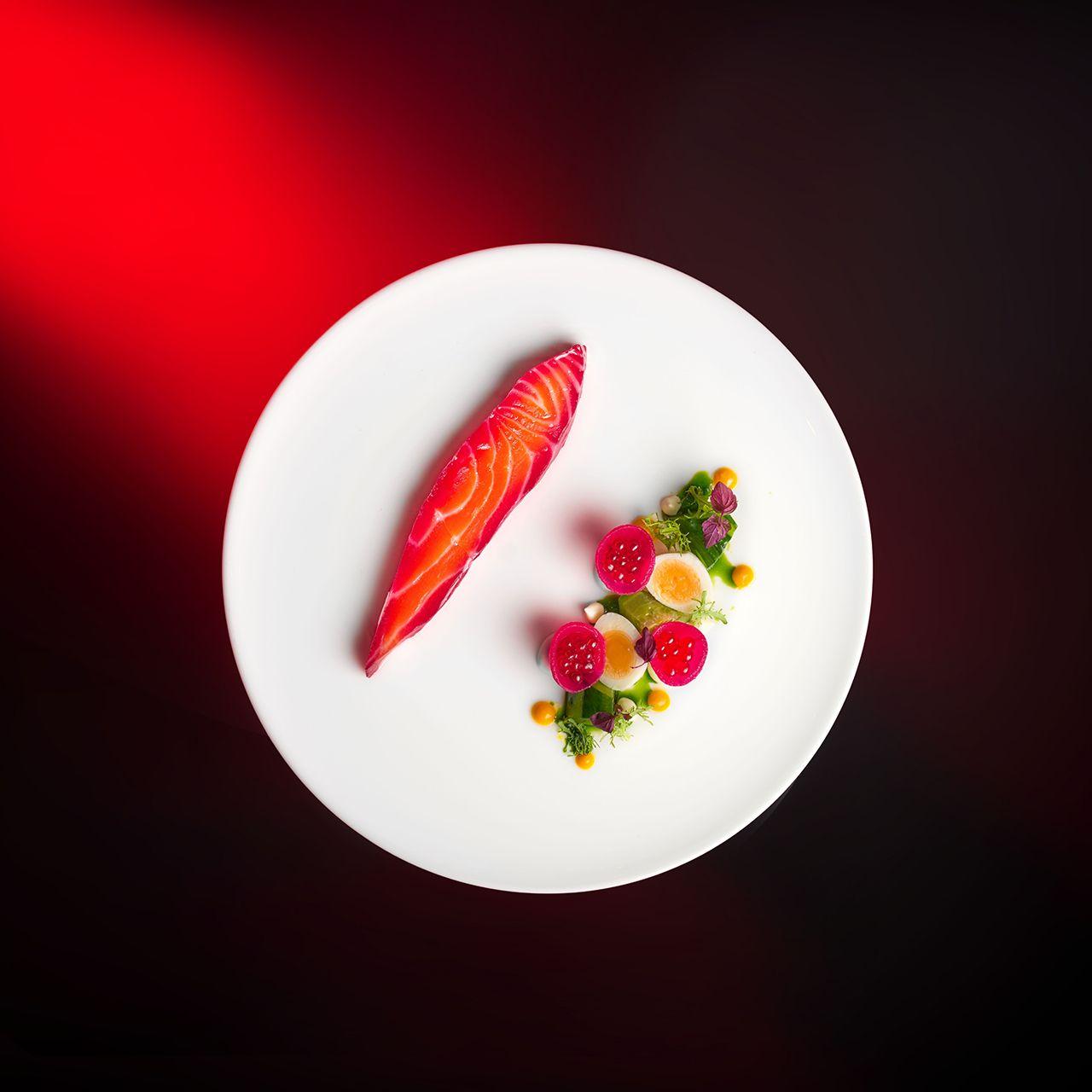 Elegantly plated cured salmon fillet on a white round plate against a dark background with red gradient. The vibrant coral-orange salmon is accompanied by an artful arrangement of thinly sliced radishes, edible flowers, microgreens, and yellow sauce dots, creating a refined and colorful composition.