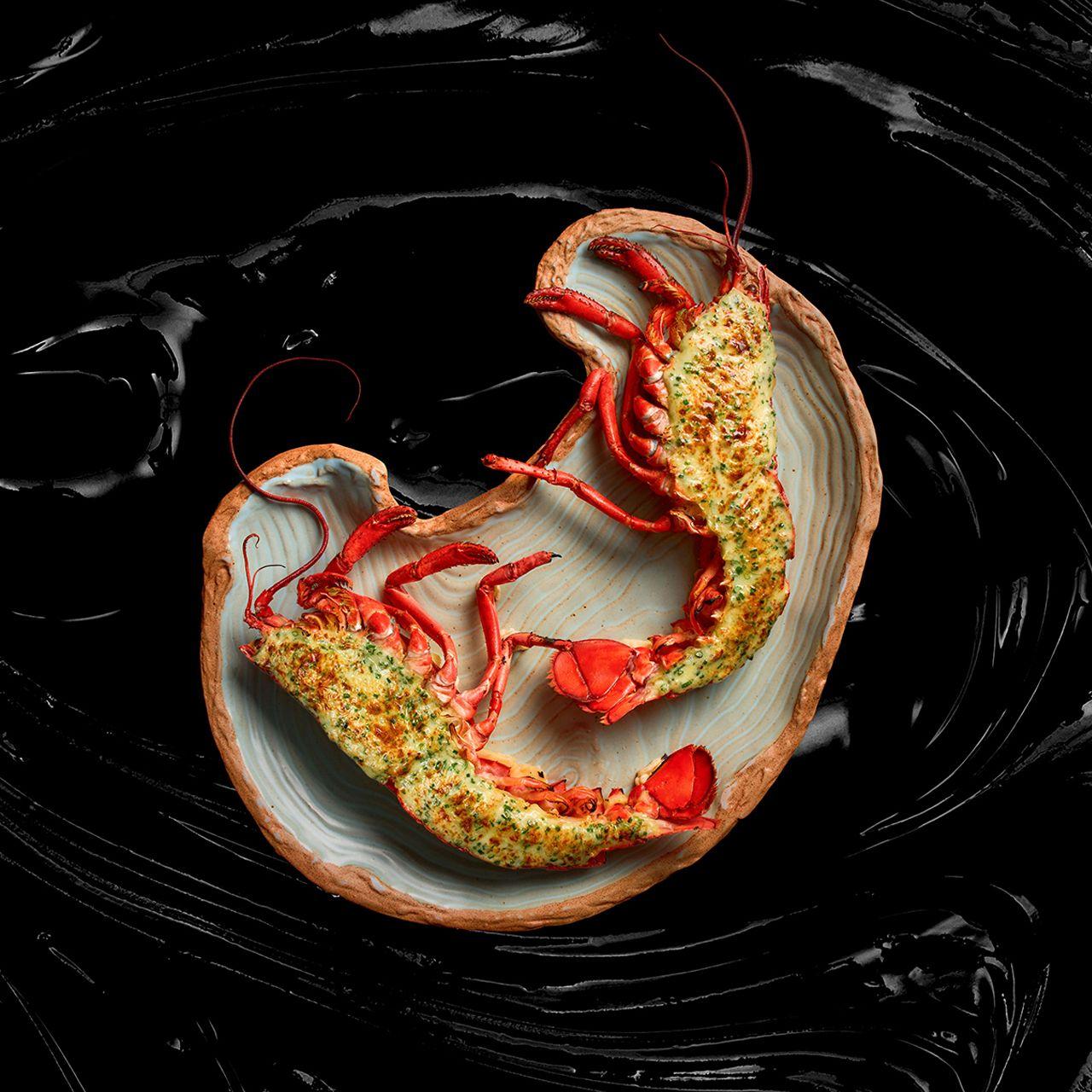 Whole lobster split and topped with herb butter gratin, served on an organic-shaped ceramic plate against a dark background.