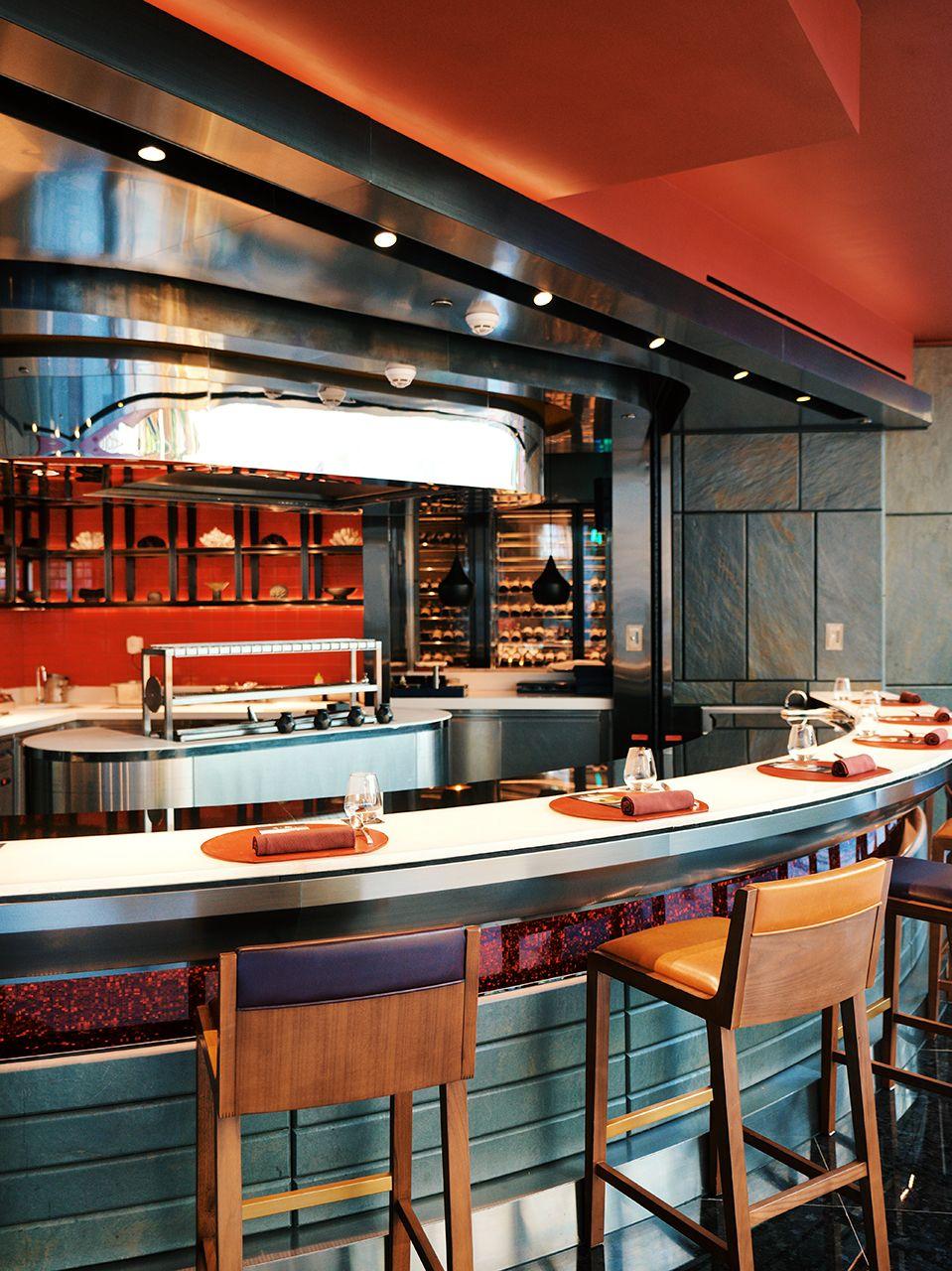Aji's open kitchen counter dining area featuring a curved white marble counter with wooden bar stools, a stainless steel hood, and red-lit ingredient display shelving in the background.