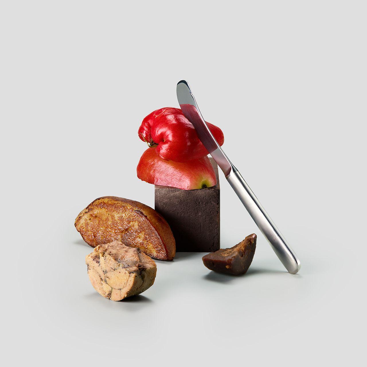 Artistic presentation featuring a glossy red pepper-shaped creation with a silver knife, accompanied by fig-like pastries on a light grey background.