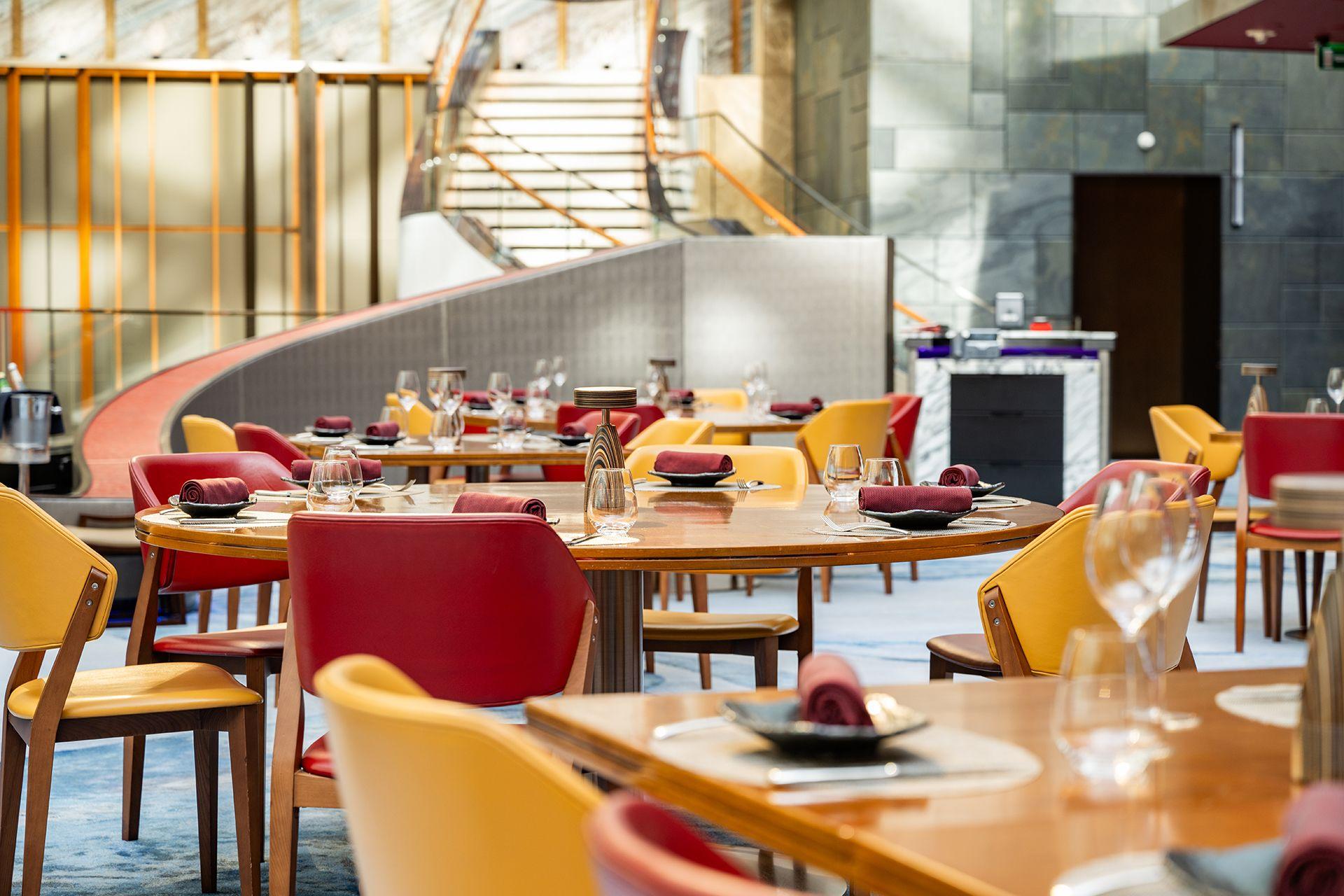 Aji restaurant's main dining area at MGM COTAI with elegantly set tables featuring burgundy napkins, yellow and red upholstered chairs, and a grand marble staircase in the background.