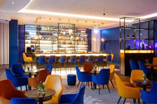 Eine Hotelbar in Berlin mit hohen Barhockern in Blau und Gelb, mehreren Tischen mit Stühlen sowie dekorativen Regalen und einer indirekten Beleuchtung.