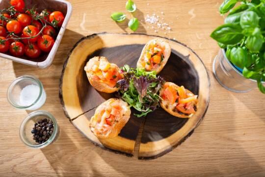 Vier Bruschetta mit Tomaten und Kräutern auf einer rustikalen Holzplatte, daneben frische Tomaten, Pfeffer, Salz und Basilikum.