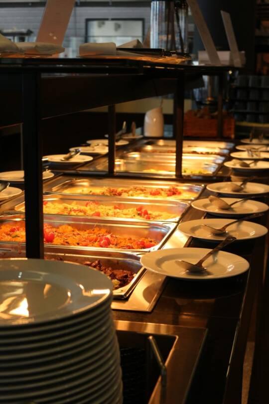 Ein Buffet mit mehreren warmen Speisen in Edelstahlbehältern und einem Stapel weißer Teller mit Besteck auf der Seite.