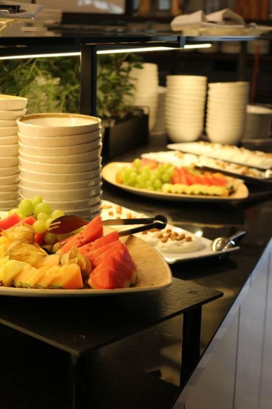 Ein Buffet mit Tellern und mehreren Platten mit geschnittenem Obst wie Melone, Ananas und Trauben auf einer schwarzen Oberfläche.