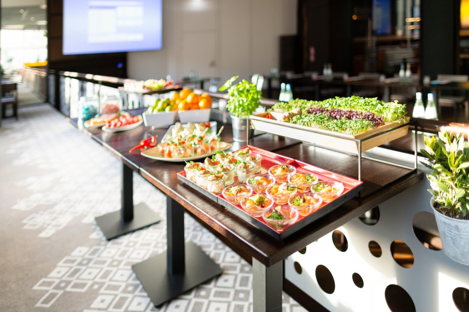 Ein Buffet vom Catering mit verschiedenen kleinen Speisen und frischen Kräutern auf einem langen Tisch in einem modernen Raum.