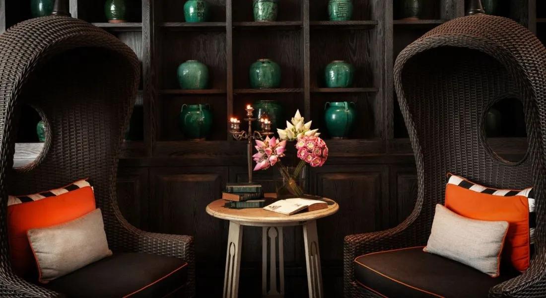 Cozy reading corner at Shinta Mani Angkor with wicker high-back chairs, lotus flowers, and green ceramic jars on display shelves.