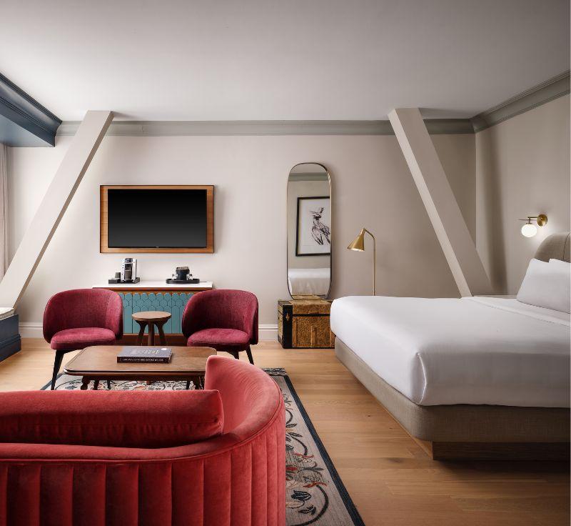 A spacious and modern hotel room with a large bed, red armchairs, and a wall-mounted flat-screen TV.